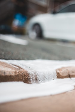A close-up view of white foam on a concrete surface, with a blurred background featuring part of a white car. The focus is on the texture and flow of the foam.