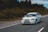 A white sports car is driving on a smooth, empty road beside a dense forest. The car has a sticker on the windshield and a sporty design.