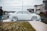 A person is washing a white car with soap using a pressure washer outside on a residential street. The car is covered in white foam, and the scene includes a grassy area with small white flowers. Several houses and parked cars are visible in the background under a cloudy sky.
