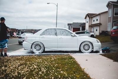A person is washing a white car with soap using a pressure washer outside on a residential street. The car is covered in white foam, and the scene includes a grassy area with small white flowers. Several houses and parked cars are visible in the background under a cloudy sky.