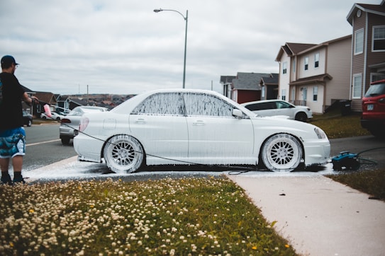 A person is washing a white car with soap using a pressure washer outside on a residential street. The car is covered in white foam, and the scene includes a grassy area with small white flowers. Several houses and parked cars are visible in the background under a cloudy sky.