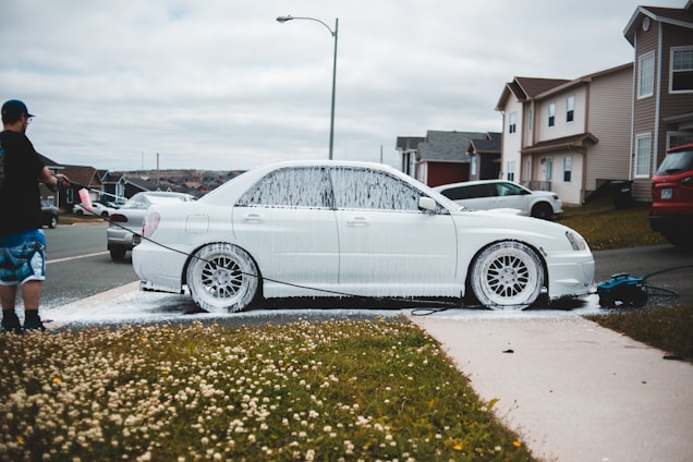 A person is washing a white car with soap using a pressure washer outside on a residential street. The car is covered in white foam, and the scene includes a grassy area with small white flowers. Several houses and parked cars are visible in the background under a cloudy sky.