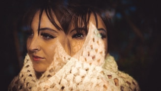 A woman is wrapped in a beige, knitted shawl with a double exposure effect, creating a surreal visual of two overlapping faces. The background is dimly lit, with hints of greenery.