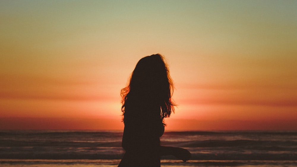 silhouette of woman standing on seashore during sunset