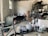 A cluttered workshop or storage room with various items scattered around. The room includes a bicycle, several paint buckets, bags, and a fire extinguisher. There are metal shelves on the right side containing tools, cans, and other materials. The walls are worn and the floor is dusty, creating a rustic atmosphere. Sunlight filters through a small window, illuminating parts of the space.