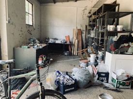 A cluttered workshop or storage room with various items scattered around. The room includes a bicycle, several paint buckets, bags, and a fire extinguisher. There are metal shelves on the right side containing tools, cans, and other materials. The walls are worn and the floor is dusty, creating a rustic atmosphere. Sunlight filters through a small window, illuminating parts of the space.