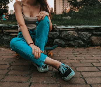 A person is sitting on a skateboard on a brick path, with legs crossed and leaning back slightly. They are wearing a tank top, blue jeans, and black sneakers. The background consists of a stone wall, grass, and blurred urban buildings. The scene has a casual and relaxed atmosphere.