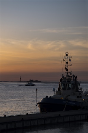 A tugboat assisting a massive vessel through calm harbor waters at sunset.