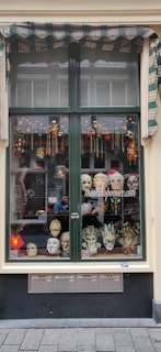 A shop window displaying an array of decorative masks, including theatrical and Venetian styles. The window has a striped awning and is bordered with a green frame. Various colorful and intricate masks are arranged on shelves, with some adorned with feathers and others with exaggerated features.