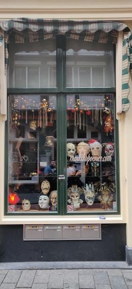 A shop window displaying an array of decorative masks, including theatrical and Venetian styles. The window has a striped awning and is bordered with a green frame. Various colorful and intricate masks are arranged on shelves, with some adorned with feathers and others with exaggerated features.