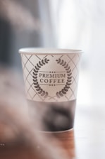 A paper cup with a decorative design featuring a laurel wreath encircling the text 'Premium Coffee'. The cup is placed on a wooden surface with soft lighting contributing to a serene and cozy atmosphere.