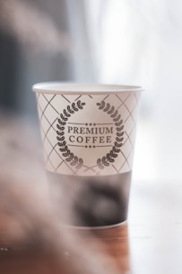 A paper cup with a decorative design featuring a laurel wreath encircling the text 'Premium Coffee'. The cup is placed on a wooden surface with soft lighting contributing to a serene and cozy atmosphere.