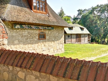A rustic scene featuring a stone cottage with a thatched roof and wooden window. Adjacent is a wooden barn with a green metal roof, surrounded by trees and grassy area. A stone wall with a tiled top runs in front.