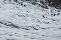 A group of people wearing colorful jackets and hiking gear is trekking across a vast snow-covered glacier. The landscape is dominated by icy, rugged terrain with patches of rock and ice.