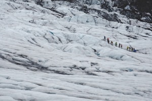 A group of people wearing colorful jackets and hiking gear is trekking across a vast snow-covered glacier. The landscape is dominated by icy, rugged terrain with patches of rock and ice.