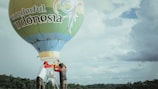 Two people are preparing a hot air balloon with 'wonderful Indonesia' written on it. The balloon is set against a backdrop of a cloudy sky and some distant foliage.