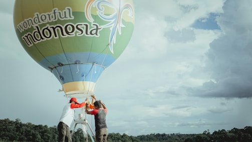 Two people are preparing a hot air balloon with 'wonderful Indonesia' written on it. The balloon is set against a backdrop of a cloudy sky and some distant foliage.
