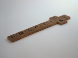 Wooden Scrabble tiles spell out the word 'PRACTISE' placed horizontally on a white surface. Each tile displays a letter and a corresponding number indicating its point value.