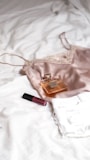 A silk camisole, a bottle of perfume, and a tube of lipstick are placed on a white textured surface, likely a bed. The camisole is made of a soft, shiny fabric with lace details, and the perfume bottle is rectangular with a golden hue. The lipstick is a deep red color.