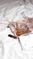 A silk camisole, a bottle of perfume, and a tube of lipstick are placed on a white textured surface, likely a bed. The camisole is made of a soft, shiny fabric with lace details, and the perfume bottle is rectangular with a golden hue. The lipstick is a deep red color.