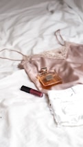 A silk camisole, a bottle of perfume, and a tube of lipstick are placed on a white textured surface, likely a bed. The camisole is made of a soft, shiny fabric with lace details, and the perfume bottle is rectangular with a golden hue. The lipstick is a deep red color.