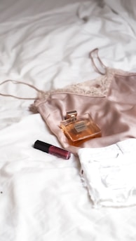 A silk camisole, a bottle of perfume, and a tube of lipstick are placed on a white textured surface, likely a bed. The camisole is made of a soft, shiny fabric with lace details, and the perfume bottle is rectangular with a golden hue. The lipstick is a deep red color.
