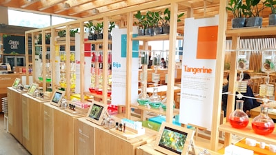 A vibrant and modern display area with wooden shelves featuring colorful glass flasks filled with various liquids. Each section is labeled with different themes like 'Tangerine' and 'Bija'. Small plants are placed on top of the shelves, and digital screens or tablets show images related to the themes. The background includes a cafe area with tables and people.