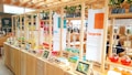 A vibrant and modern display area with wooden shelves featuring colorful glass flasks filled with various liquids. Each section is labeled with different themes like 'Tangerine' and 'Bija'. Small plants are placed on top of the shelves, and digital screens or tablets show images related to the themes. The background includes a cafe area with tables and people.