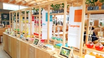 A vibrant and modern display area with wooden shelves featuring colorful glass flasks filled with various liquids. Each section is labeled with different themes like 'Tangerine' and 'Bija'. Small plants are placed on top of the shelves, and digital screens or tablets show images related to the themes. The background includes a cafe area with tables and people.