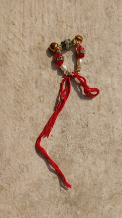 A decorative piece composed of beads and small bells arranged in a circular pattern, with a notable red thread tied onto it, lying on a textured, beige surface.