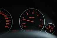 A close-up view of a car's dashboard displaying various gauges and dials with glowing orange lights. The tachometer is prominently featured in the center, surrounded by other indicators like temperature and oil pressure.