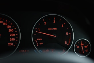 A close-up view of a car's dashboard displaying various gauges and dials with glowing orange lights. The tachometer is prominently featured in the center, surrounded by other indicators like temperature and oil pressure.