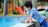 A young child wearing a blue striped shirt leans over the edge of a swimming pool, touching the water. In the background, there is a colorful playground with a slide and trees.