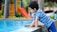 A young child wearing a blue striped shirt leans over the edge of a swimming pool, touching the water. In the background, there is a colorful playground with a slide and trees.