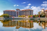A riverside view of a multi-story building with symmetrical architecture and a red-tiled roof, reflected vividly in the calm water. The scene is framed by lush green trees on the banks, under a clear blue sky dotted with a few clouds. A prominent hashtag signage on the riverbank suggests a popular location.