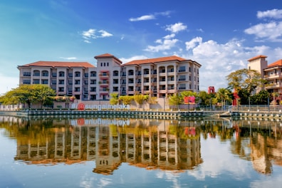 A riverside view of a multi-story building with symmetrical architecture and a red-tiled roof, reflected vividly in the calm water. The scene is framed by lush green trees on the banks, under a clear blue sky dotted with a few clouds. A prominent hashtag signage on the riverbank suggests a popular location.