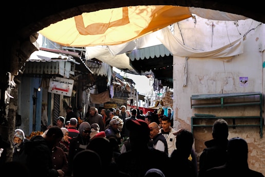 A bustling marketplace scene with a crowd of people engaged in various activities under a canopy. Stalls and shops are visible on either side, and the architecture features weathered, aged walls.