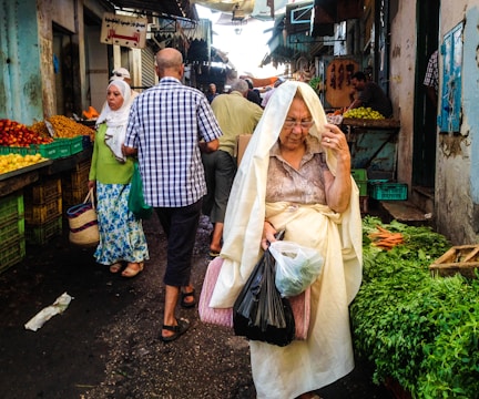 A bustling market scene with several people walking between stalls. An older woman wrapped in a light-colored cloth is carrying plastic bags filled with produce. Other people, including a woman in a white headscarf and a man in a checked shirt, are also visible. Various fruits and vegetables are displayed in crates, including herbs and carrots.