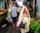 A bustling market scene with several people walking between stalls. An older woman wrapped in a light-colored cloth is carrying plastic bags filled with produce. Other people, including a woman in a white headscarf and a man in a checked shirt, are also visible. Various fruits and vegetables are displayed in crates, including herbs and carrots.
