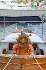 A wooden boat with a detailed steering wheel is docked at a marina. The boat has a canopy overhead and life buoys are visible near the helm. The background shows another boat and a coastal town with several buildings.