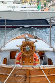 A wooden boat with a detailed steering wheel is docked at a marina. The boat has a canopy overhead and life buoys are visible near the helm. The background shows another boat and a coastal town with several buildings.