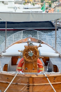 A wooden boat with a detailed steering wheel is docked at a marina. The boat has a canopy overhead and life buoys are visible near the helm. The background shows another boat and a coastal town with several buildings.