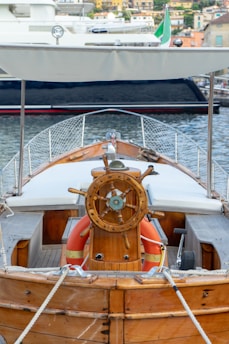 A wooden boat with a detailed steering wheel is docked at a marina. The boat has a canopy overhead and life buoys are visible near the helm. The background shows another boat and a coastal town with several buildings.