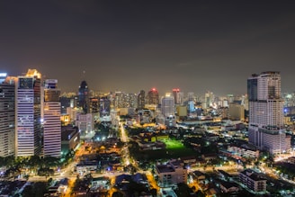 A vibrant cityscape at night featuring numerous illuminated skyscrapers and buildings. Streets are bustling with activity, and the city lights create a warm glow against the dark sky. The area is densely populated with a variety of architectural styles.