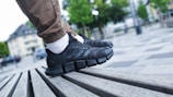 A close-up shot of a person's feet wearing black athletic shoes with distinctive thick, cushioned soles. The person is standing on a slatted wooden bench in an outdoor urban setting. The individual is wearing light brown pants and white socks. The background shows an out-of-focus park area with trees and tall buildings.