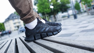 A close-up shot of a person's feet wearing black athletic shoes with distinctive thick, cushioned soles. The person is standing on a slatted wooden bench in an outdoor urban setting. The individual is wearing light brown pants and white socks. The background shows an out-of-focus park area with trees and tall buildings.