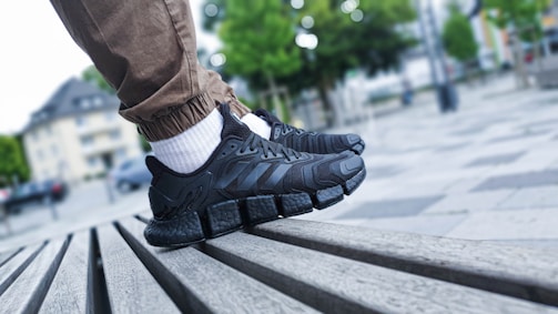 A close-up shot of a person's feet wearing black athletic shoes with distinctive thick, cushioned soles. The person is standing on a slatted wooden bench in an outdoor urban setting. The individual is wearing light brown pants and white socks. The background shows an out-of-focus park area with trees and tall buildings.