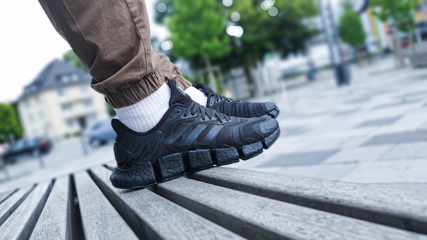 A close-up shot of a person's feet wearing black athletic shoes with distinctive thick, cushioned soles. The person is standing on a slatted wooden bench in an outdoor urban setting. The individual is wearing light brown pants and white socks. The background shows an out-of-focus park area with trees and tall buildings.