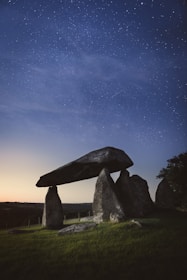 A megalithic structure consisting of large standing stones supporting a massive horizontal slab sits in an open field under a clear, star-filled night sky. Surrounding the structure is a lush landscape with gentle hills and scattered trees.