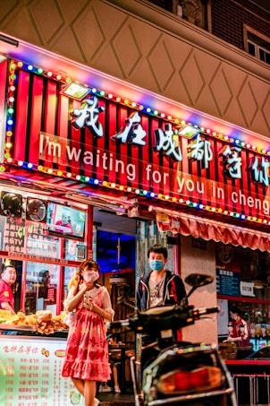 A vibrant street scene in front of a brightly lit store with a marquee sign that reads 'I'm waiting for you in Chengdu'. The store is decorated with colorful lights and Chinese characters. A man and a woman wearing face masks stand outside the entrance, with a table displaying various snacks and a scooter partially visible in the foreground. Another person is seen in the background behind the counter.